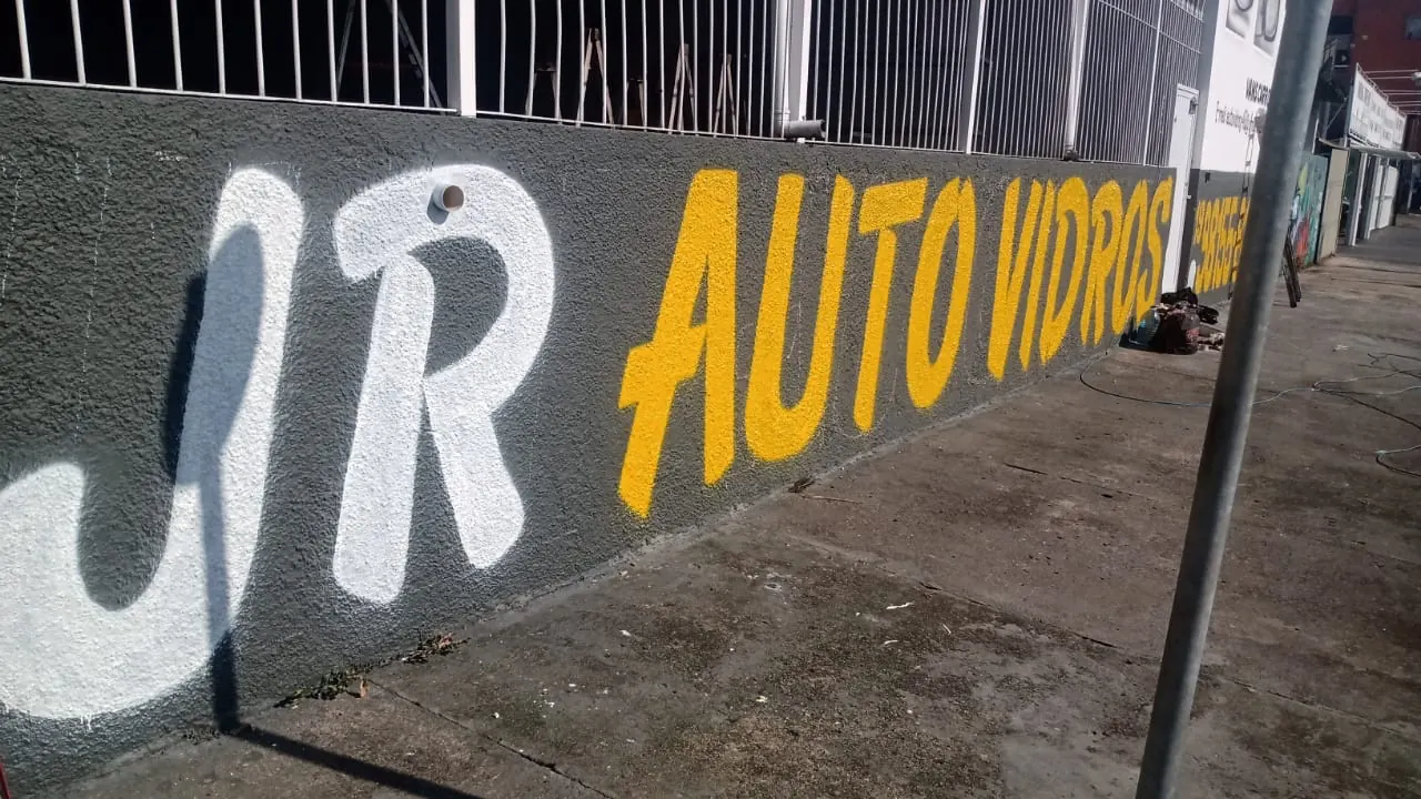 Muro da oficina pintado à mão com JR em branco e Auto Vidros em amarelo sobre cinza grafite
